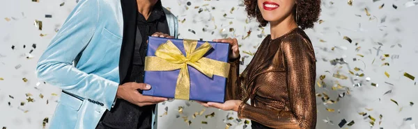 Abgeschnittene Ansicht einer lächelnden afrikanisch-amerikanischen Frau und eines eleganten Mannes mit Weihnachtsgeschenk auf grauem Hintergrund, Banner — Stockfoto