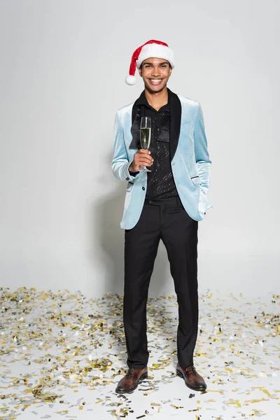 Cheerful african american man in silk blazer and santa hat standing with champagne glass on confetti on grey background — Stock Photo
