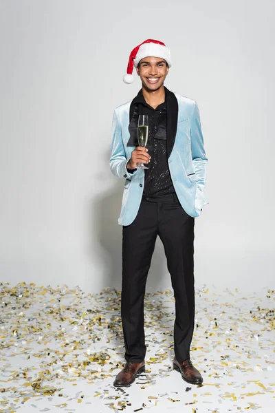 Hombre americano africano alegre en chaqueta de seda y sombrero de santa de pie con copa de champán en confeti sobre fondo gris - foto de stock