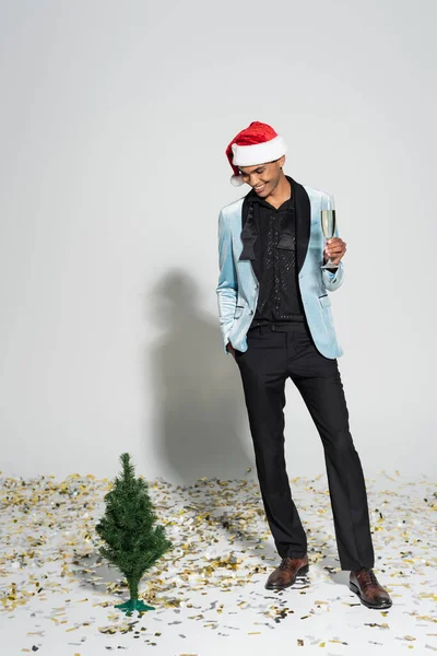 African american man in elegant clothes and santa hat holding champagne near small christmas tree on grey — Stock Photo