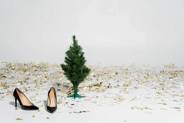Sapatos elegantes e pretos de salto alto perto de árvore de natal verde e confete espumante no fundo cinza — Fotografia de Stock