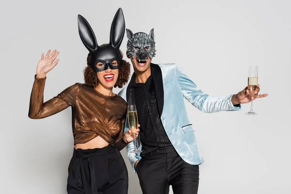 Cheerful woman in bunny mask waving hand near man in wolf mask on christmas party isolated on grey — Stock Photo