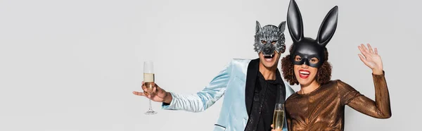 Excitada pareja afroamericana en máscaras de lobo y conejo sosteniendo champán aislado en gris, bandera - foto de stock