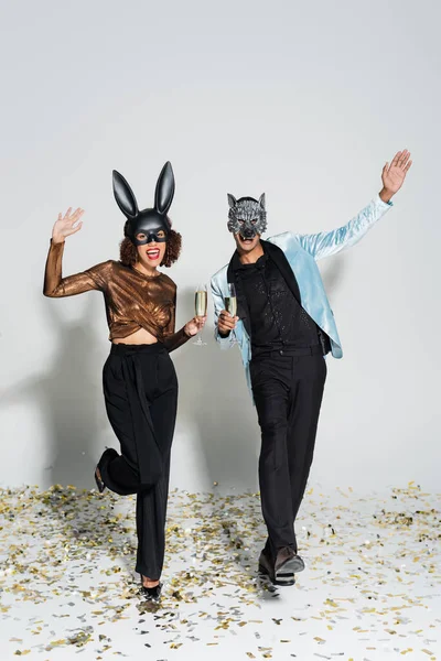 Animado casal afro-americano em coelho e lobo máscaras dançando com champanhe e acenando as mãos sobre fundo cinza — Fotografia de Stock