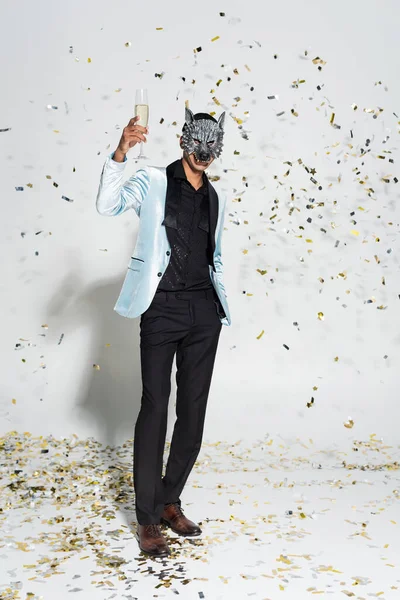 Full length of stylish african american man in wolf mask toasting with champagne near confetti on grey background — Stock Photo