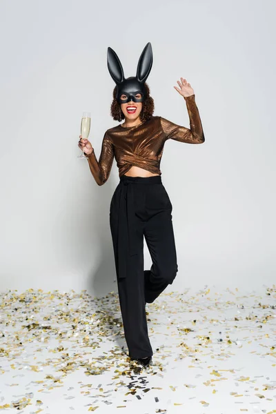 Cheerful african american woman in elegant clothes and bunny mask holding champagne and waving hand on grey background — Stock Photo