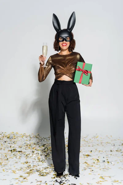Mulher americana africana elegante em máscara de coelho segurando champanhe e Natal presente no fundo cinza — Fotografia de Stock