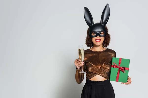 Афроамериканська жінка в елегантному одязі і масках кролика стоїть з шампанським і зеленим подарунковим ящиком на сірому фоні — стокове фото