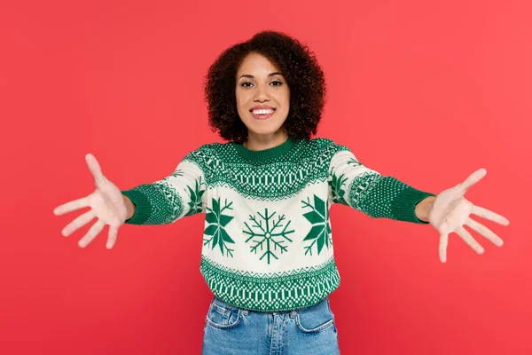 Mujer afroamericana feliz en suéter de invierno con patrón verde mirando a la cámara con las manos extendidas aisladas en rojo - foto de stock