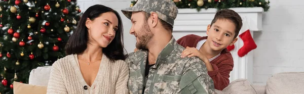 Homem militar com esposa feliz olhando uns para os outros perto do filho e árvore de natal decorada, bandeira — Fotografia de Stock