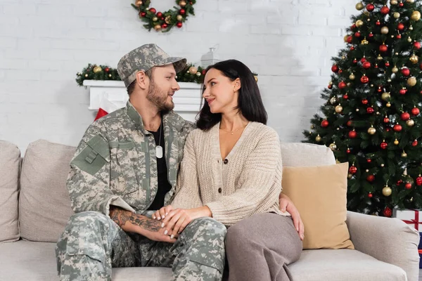 Homem em camuflagem abraçando esposa feliz enquanto sentado no sofá perto da árvore de natal decorada — Fotografia de Stock