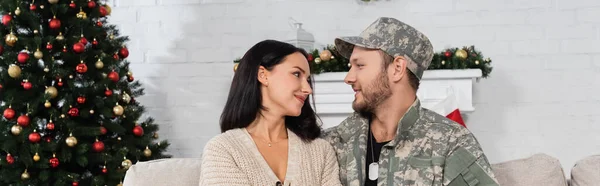 Happy woman looking at husband in military uniform in living room with christmas tree, banner — Stock Photo