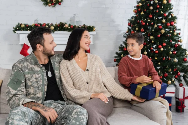 Хлопчик з різдвяним подарунком сидить на дивані біля матері і батька у військовій формі — стокове фото