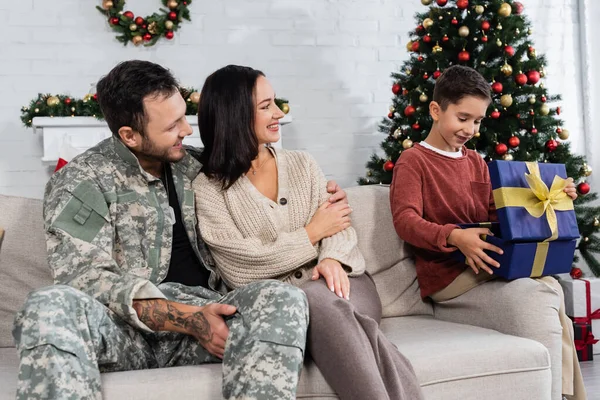 Niño sonriente abriendo caja de regalo cerca de la madre y el padre en camuflaje sentado en el sofá - foto de stock