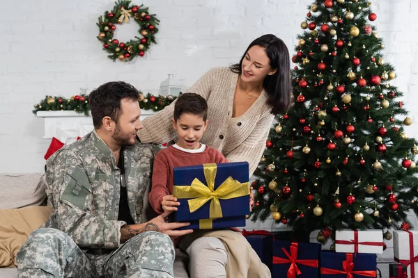 Niño sorprendido abriendo caja de regalo cerca del padre en camuflaje y madre sonriente en la sala de estar con árbol de Navidad decorado - foto de stock