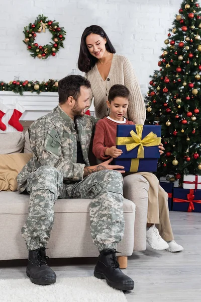 Fröhliche Frau schaut Sohn beim Öffnen der Geschenkbox neben Vater in Tarnung und geschmücktem Weihnachtsbaum an — Stockfoto