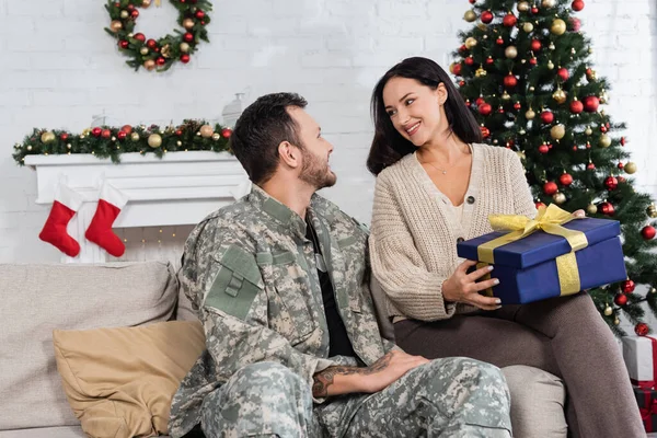 Веселая женщина с подарочной коробкой глядя на мужа в камуфляже рядом с украшенной елкой — стоковое фото