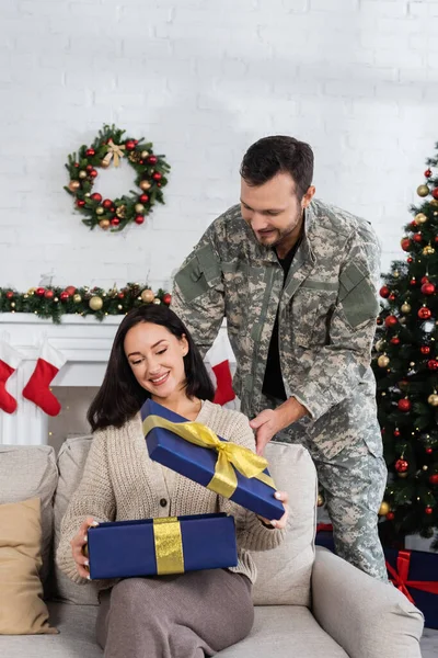 Mujer complacida abriendo caja de regalo cerca del marido en camuflaje en la sala de estar con decoración de Navidad - foto de stock