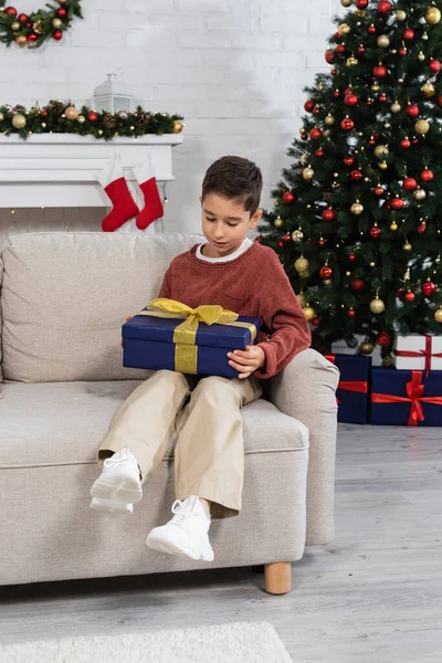 Ребенок в полный рост с подарком, сидящий на диване рядом с украшенной елкой дома — стоковое фото