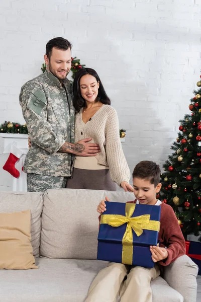 Feliz homem militar abraçando esposa perto de árvore de natal decorada e filho caixa de presente de abertura — Fotografia de Stock