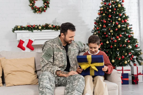Изумленный мальчик смотрит в подарочную коробку рядом с папой в камуфляже и рождественская елка с украшенным камином — стоковое фото