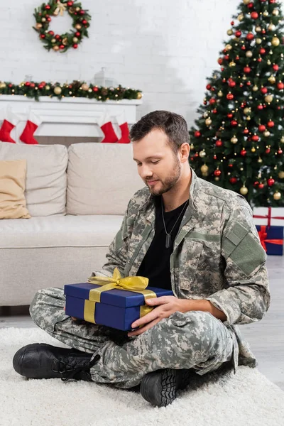 Homme militaire souriant assis sur le sol avec boîte cadeau près de l'arbre de Noël et cheminée décorée — Photo de stock