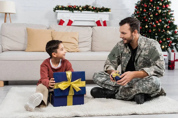 Niño feliz y papá en camuflaje sentado en la alfombra del piso con cajas de regalo en la sala de estar decorada para Navidad - foto de stock