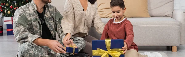 Smiling boy opening gift box near mother and dad in military uniform, banner — Stock Photo