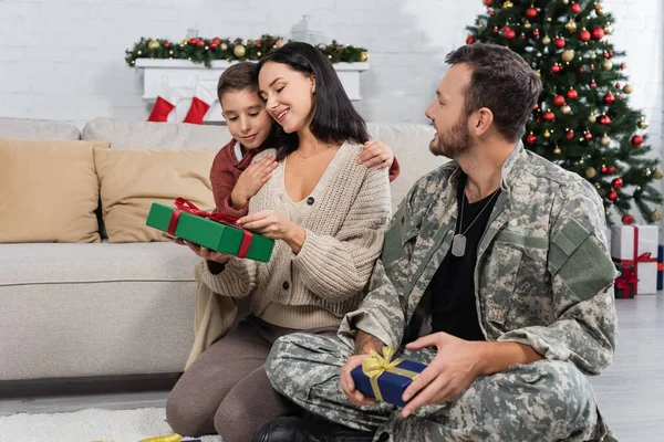 Ребенок обнимает улыбающуюся мать, сидящую на полу и держащую рождественский подарок рядом с мужем в камуфляже — стоковое фото