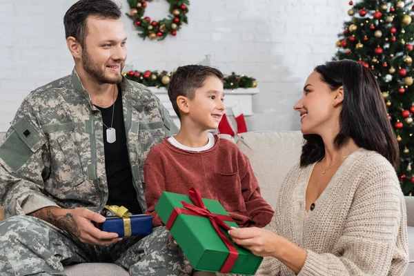 Alegre mujer sosteniendo navidad presente cerca de hijo y marido en uniforme militar - foto de stock