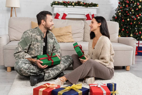 Військовий чоловік з щасливою дружиною сидить на підлозі біля подарункових коробок і посміхається один одному — стокове фото