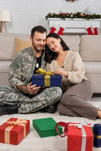 Улыбающаяся женщина, открывающая рождественский подарок, сидя на полу рядом с мужем в камуфляже — стоковое фото