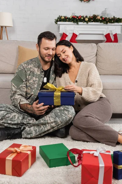 Smiling woman opening christmas present while sitting on floor near husband in camouflage — Stock Photo