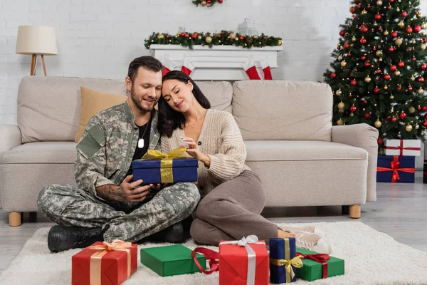 Щаслива жінка з чоловіком у військовій формі сидить на підлозі біля подарункових коробок та прикрашає ялинку — стокове фото