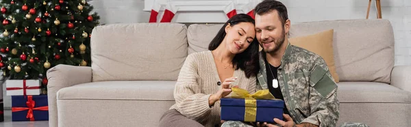 Heureuse femme ouverture Noël présent près de mari en camouflage, bannière — Photo de stock