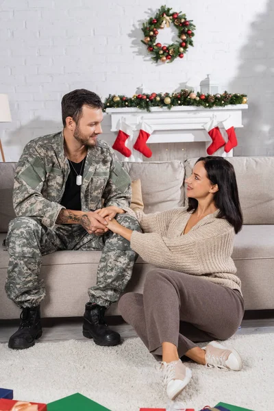 Усміхнена жінка сидить на підлозі і тримає руки чоловіка в камуфляжі у вітальні з різдвяним декором — стокове фото