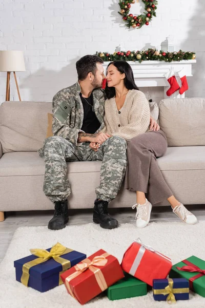Військовий чоловік сидить обличчям до обличчя з дружиною біля купи подарункових коробок у вітальні з різдвяним декором — стокове фото