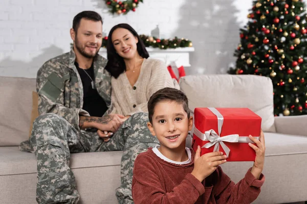 Junge mit Weihnachtsgeschenk lächelt in die Kamera neben Mutter und Vater in Tarnkleidung — Stockfoto