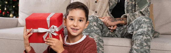 Niño feliz con regalo de Navidad mirando a la cámara cerca de mamá y papá en camuflaje cogido de la mano en el fondo, pancarta - foto de stock