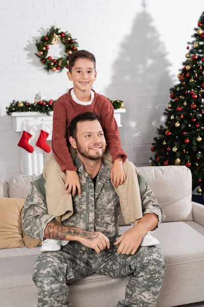 Alegre hombre en camuflaje piggybacking sonriente hijo mientras está sentado en el sofá cerca de árbol de Navidad - foto de stock