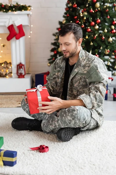 Pleased military man holding christmas present while sitting on floor carpet in decorated living room — Stock Photo