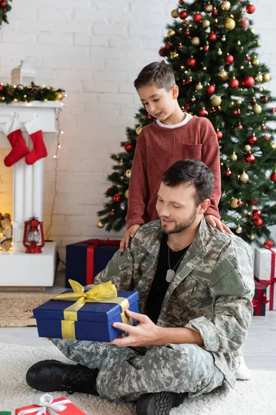 Glücklicher Soldat hält Neujahrsgeschenk in der Nähe von Sohn und verschwommenem Weihnachtsbaum — Stockfoto