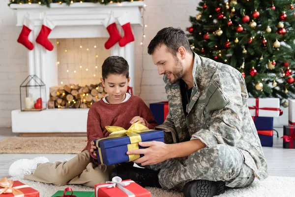 Веселий військовий чоловік сидить з подарунковою коробкою біля сина у вітальні з ялинкою та каміном — стокове фото