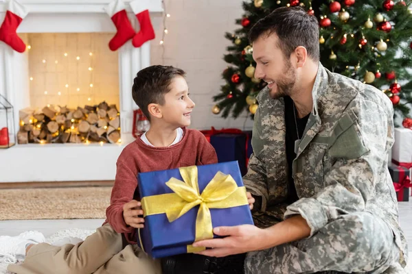 Joyful kid and father in camouflage holding gift box and smiling at each other near decorated fireplace and christmas tree — Stock Photo