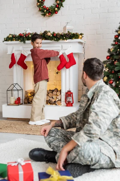 Smiling boy holding christmas stocking near fireplace and father in camouflage sitting on blurred foreground — Stock Photo