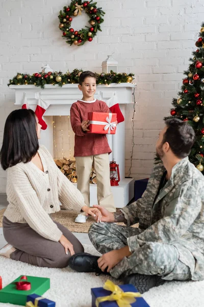 Hombre en camuflaje sentado en el suelo cerca de la esposa y el hijo feliz celebración de regalo de Navidad cerca de la chimenea decorada - foto de stock