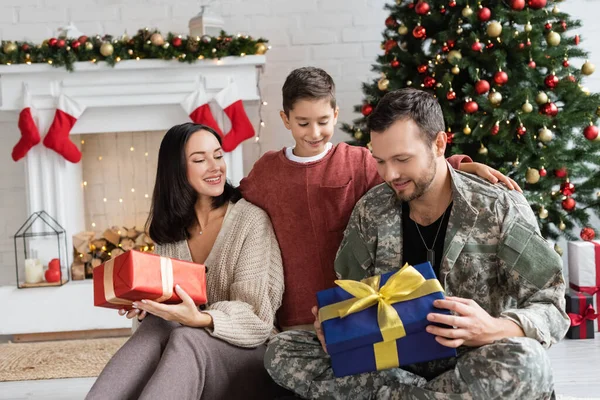 Homem militar feliz segurando caixa de presente perto de esposa e filho na sala de estar com árvore de natal — Fotografia de Stock