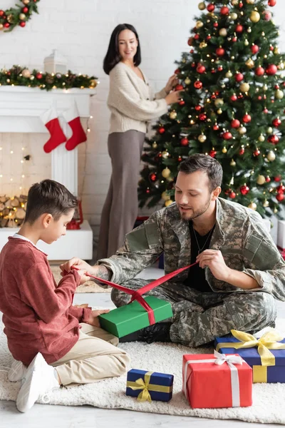 Militär verpackt Geschenkboxen mit Sohn in der Nähe Frau schmücken Weihnachtsbaum auf verschwommenem Hintergrund — Stockfoto