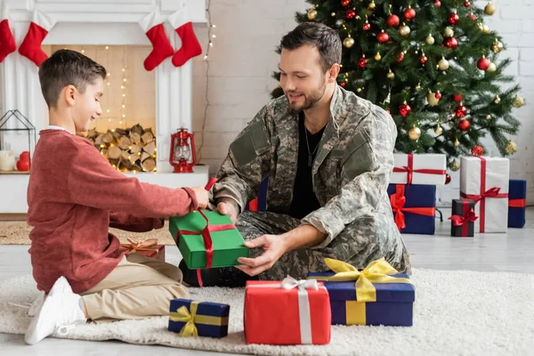 Lächelnder Mann in Tarnkleidung sitzt auf dem Boden neben Sohn, der Weihnachtsgeschenke einpackt — Stockfoto