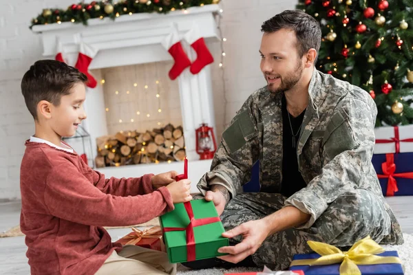 Хлопчик упаковує подарункову коробку біля батька в камуфляжі, сидячи біля каміна з різдвяним декором — стокове фото